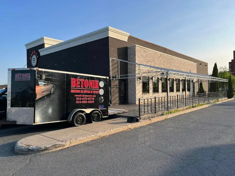 Bétonik Inc. trailer parked outside a commercial building, showcasing residential epoxy flooring services in Greater Montreal, featuring branding and contact information.