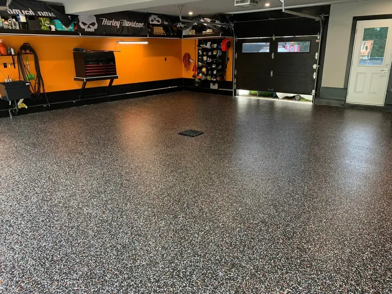 Epoxy flooring in a garage setting, featuring a smooth black and gray speckled finish, with orange walls, storage tools, and garage door visible.