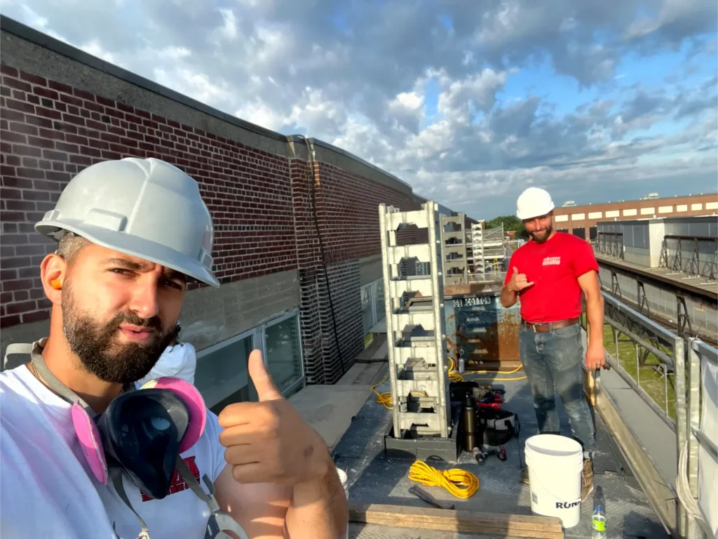 Construction workers on a rooftop, one giving a thumbs-up, wearing safety helmets and gear, with tools and materials for epoxy and concrete work visible in the background.