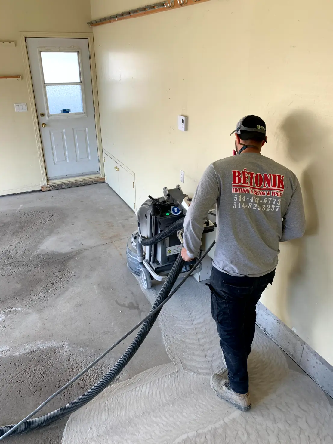 Worker operating a concrete grinder in a garage, preparing the floor for epoxy installation, wearing a Bétonik branded shirt.