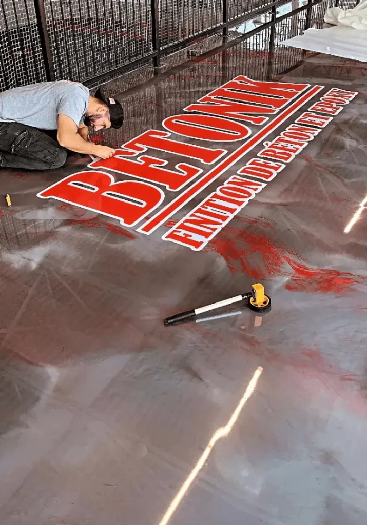 Travailleur appliquant un logo Bétonik sur une surface de sol en époxy, mettant en avant la finition professionnelle et moderne pour le centre sportif de Saint-Roch.