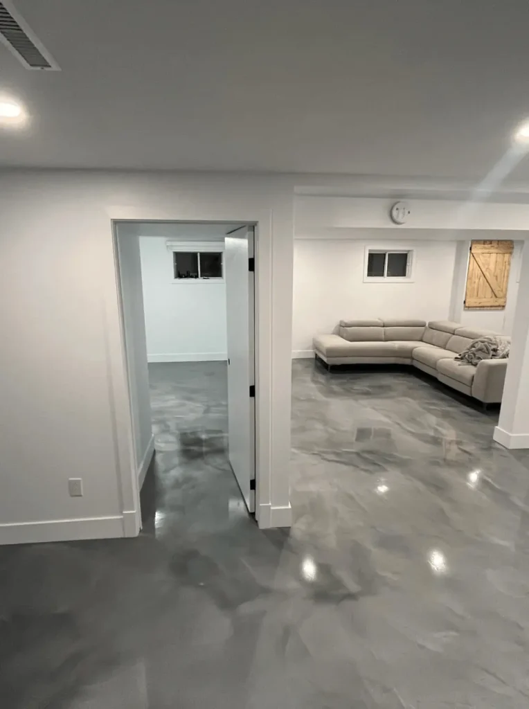 Modern basement interior featuring polished epoxy flooring, white walls, open doorway, and a gray sectional sofa, highlighting elegance and moisture protection.