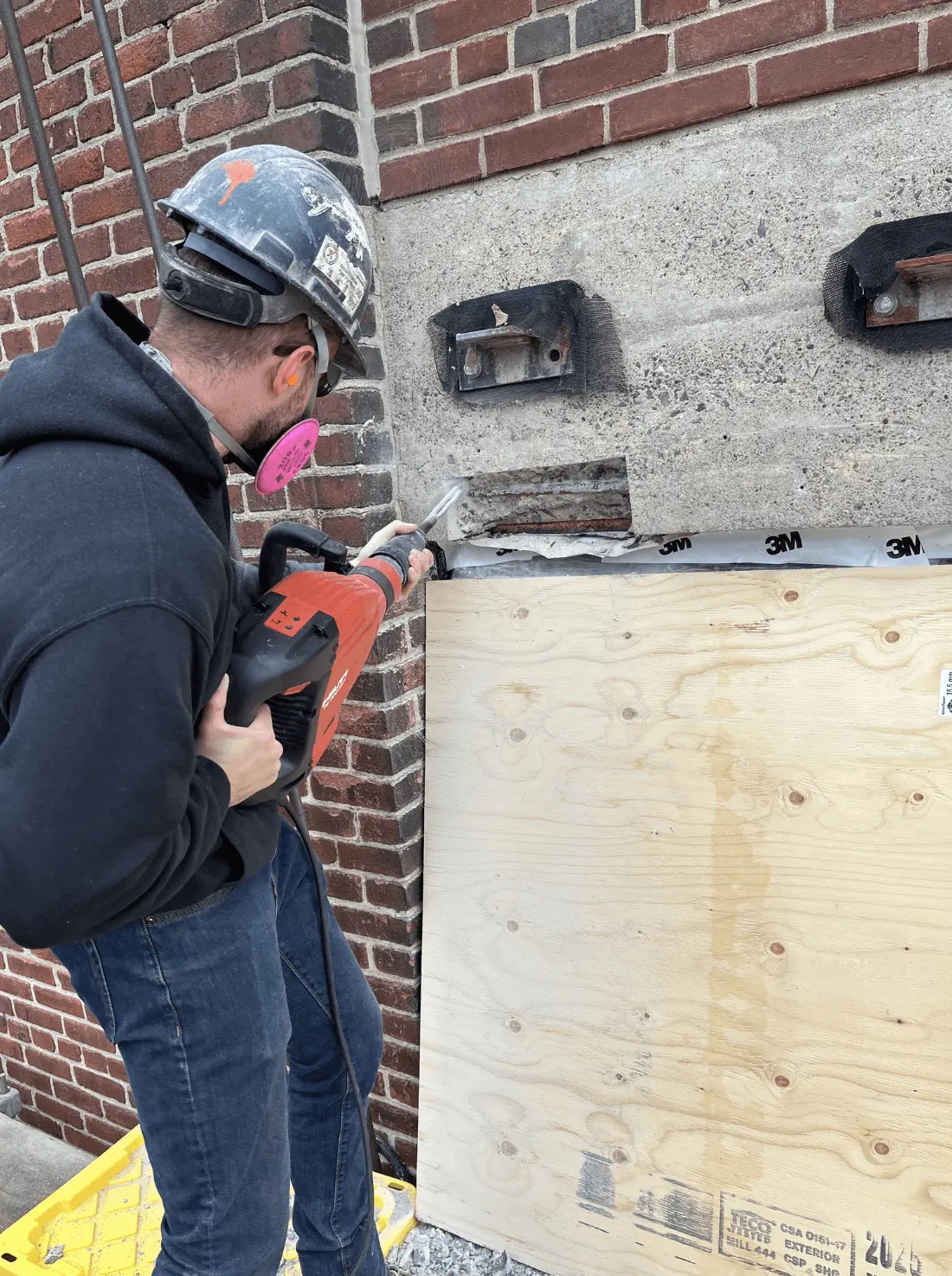 Worker using a power tool to remove deteriorated concrete from a wall, preparing for structural repair in a concrete restoration project.