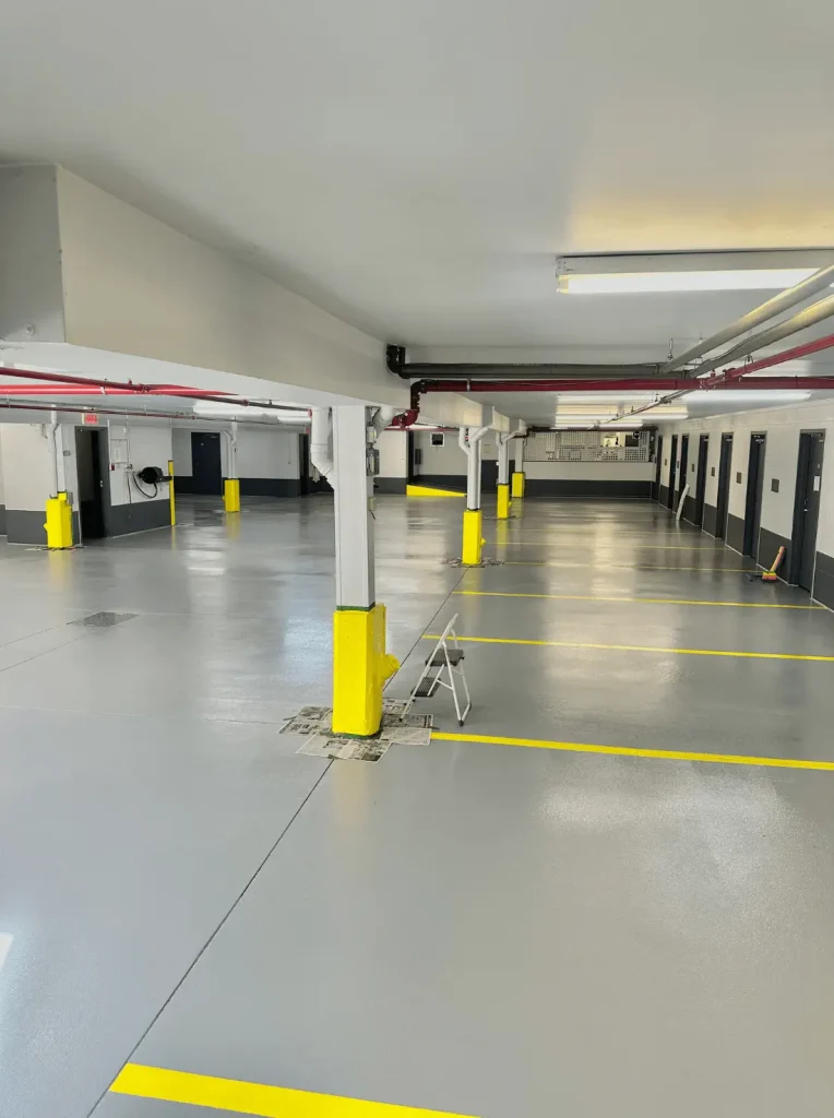 Indoor parking lot with freshly coated gray epoxy flooring, yellow-painted support columns, and clear yellow parking lines, showcasing professional-grade concrete repair and resurfacing services by Bétonik.