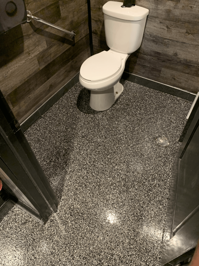 Epoxy flooring in a commercial restroom featuring a glossy black surface, complemented by wooden wall panels and a white toilet, illustrating durable and easy-to-maintain flooring suitable for restaurants.