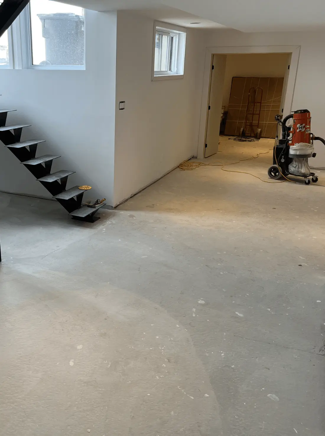 Basement interior with exposed concrete floor, metal staircase, and construction equipment, illustrating the preparation stage for epoxy flooring application by Bétonik.