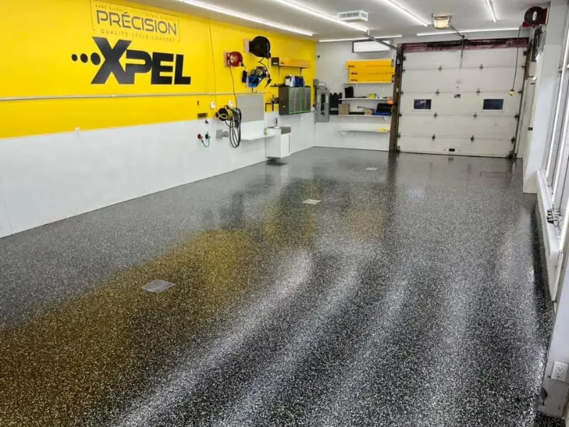 Garage interior featuring high-gloss epoxy flooring with a custom flake blend, yellow wall with "PRÉCISION" and "...XPEL" branding, and organized tools and equipment, illustrating the professional results of Bétonik's epoxy flooring services for automotive detailing.