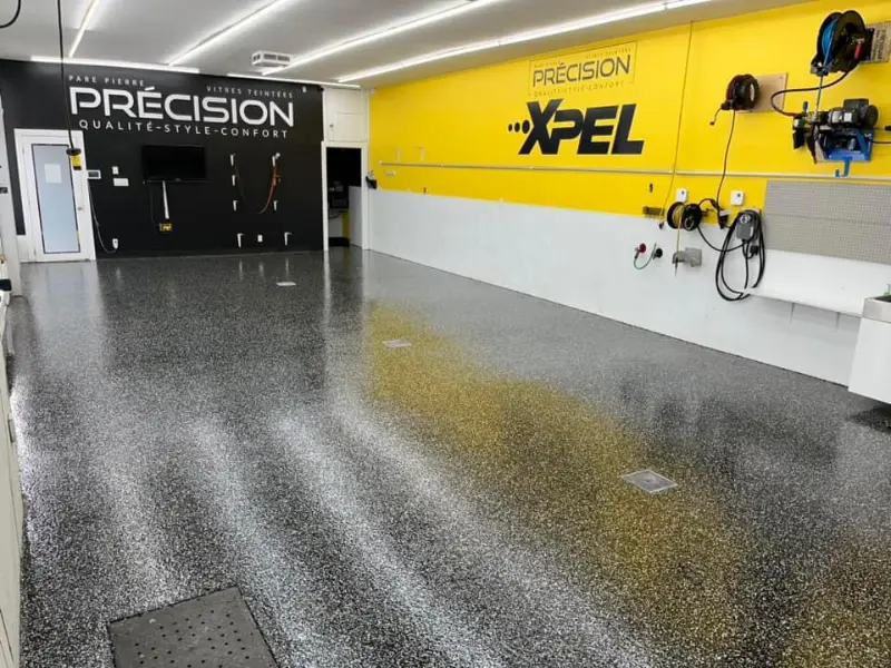 Salle de détail automobile avec sol en époxy brillant, logo "PRÉCISION" sur mur noir, et "XPEL" sur mur jaune, démontrant l'esthétique premium et la durabilité du revêtement.