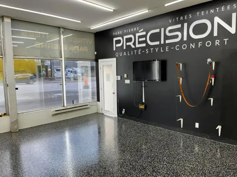 Intérieur d'un centre de détail automobile avec un sol en époxy brillant, affichant le logo "PRÉCISION" sur un mur noir, fenêtres laissant entrer la lumière, et équipements muraux visibles, illustrant l'esthétique premium et la durabilité des services de Bétonik.
