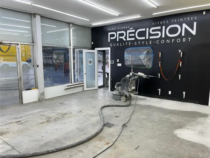 Intérieur d'un centre de detailing automobile avec un mur noir affichant "PRÉCISION QUALITÉ-STYLE-CONFORT", équipement de préparation de sol en cours d'utilisation, et espace prêt pour l'application de revêtement époxy.