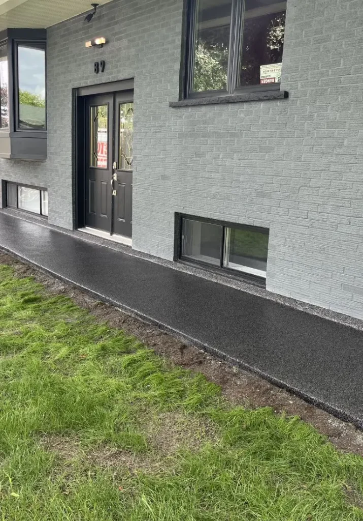 Finition en béton noir autour de l'entrée principale d'une maison, avec pelouse verte et fenêtres modernes, illustrant les services de Bétonik pour des aménagements extérieurs élégants et durables.