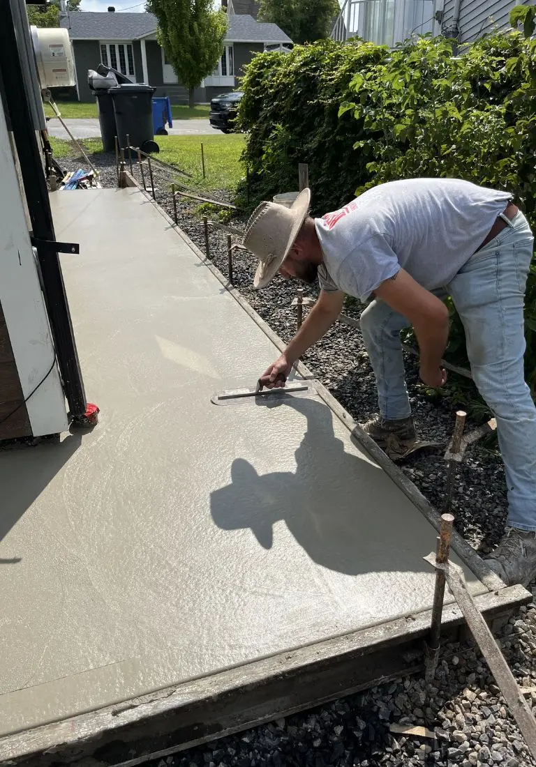 Homme appliquant du béton frais sur un trottoir, utilisant une truelle pour obtenir une finition lisse, entouré de coffrages et de barres d'armature.